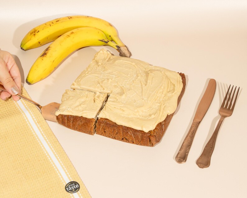 May include: A square piece of banana bread topped with a thick layer of vanilla frosting. Two ripe bananas and a butter knife are on the table next to the bread. Two gold forks are on the table to the right of the bread.