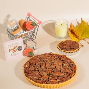 May include: A pecan pie with a golden crust and a generous amount of pecans. The pie is on a beige surface with a small pecan pie, a shopping trolley with a jar of spices, two peaches, and a candle in the background. The text "Happy Bakery Pecan Pie" is on a pink card.