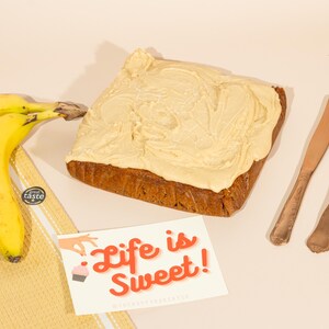May include: A frosted banana cake with a light brown frosting and the text "Life is Sweet!" on a card next to it. There are two gold forks and a gold knife on a light brown surface.