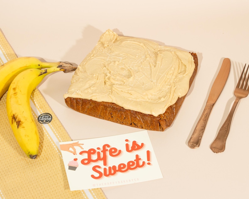 May include: A frosted banana cake with a light brown frosting and the text "Life is Sweet!" on a card next to it. There are two gold forks and a gold knife on a light brown surface.