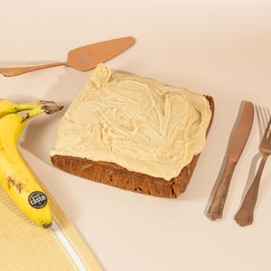 May include: A square banana cake with a swirl of light brown frosting on top. The cake is on a light brown surface with two bananas, a silver fork, a silver knife, and a copper cake server.