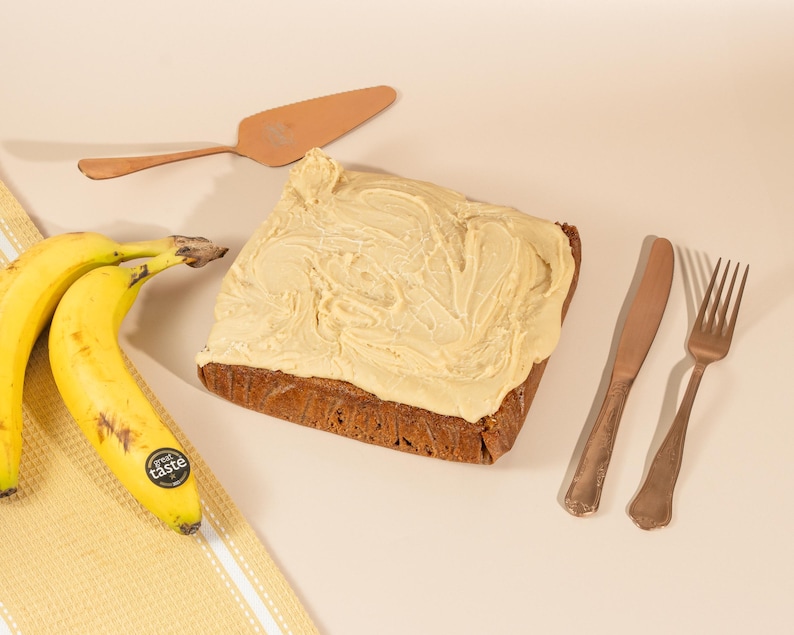 May include: A square banana cake with a swirl of light brown frosting on top. The cake is on a light brown surface with two bananas, a silver fork, a silver knife, and a copper cake server.