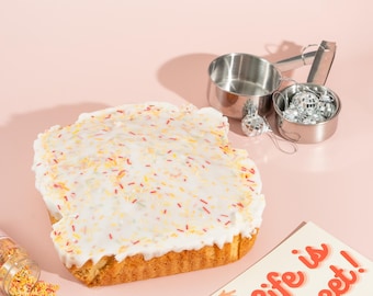 May include: A white cake with yellow and red sprinkles and white frosting. The cake is on a pink surface with a card that says "Life is Sweet!" and a small container of sprinkles.