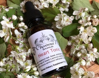 Könnte beinhalten: Eine braune Glasflasche mit einem weißen Etikett, auf dem "Reverence Botanicals Heart Tonic" steht. Das Etikett enthält auch eine Liste der Zutaten, darunter Blätter, Blüten und Beeren des Weißdorns. Die Flasche steht auf einem Bett aus weißen Blüten.