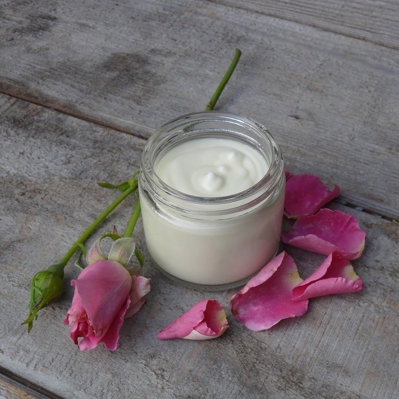 Puede incluir: Un tarro de vidrio lleno de crema blanca, rodeado de p&eacute;talos de rosa rosados y un capullo de rosa sobre una superficie de madera gris.
