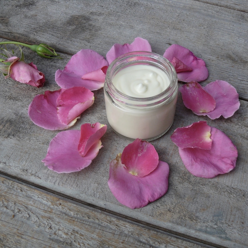 Puede incluir: Un tarro de vidrio de crema blanca rodeado de p&eacute;talos de rosa rosados sobre una superficie de madera gris.