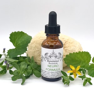 May include: A brown glass bottle with a black dropper top and a white label that reads "Reverence Botanicals Neuro Formula" with a list of ingredients including Fresh Lion's Mane mushroom, Gotu Kola, and Kudzu. The bottle is sitting on a white surface with green plants and a white sponge-like object.