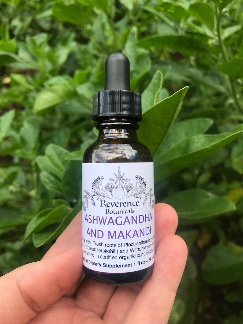 Puede incluir: Una botella de vidrio marr&oacute;n con un gotero negro y una etiqueta blanca. La etiqueta dice "Reverence Botanicals Ashwagandha and Makandi" e incluye una descripci&oacute;n de los ingredientes del producto y una etiqueta de suplemento diet&eacute;tico. La botella se sostiene en una mano contra un fondo de hojas verdes.