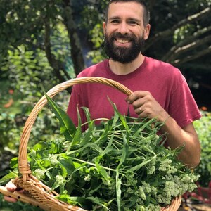 May include: A man with a beard holds a wicker basket full of fresh green leafy herbs.