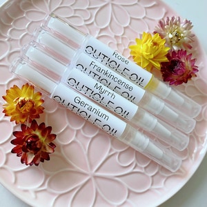 May include: Four cuticle oil pens labelled Rose, Frankincense, Myrrh, and Geranium, on a pink floral plate. The pens are clear with white labels and caps. Yellow and maroon dried flowers are scattered around the pens.