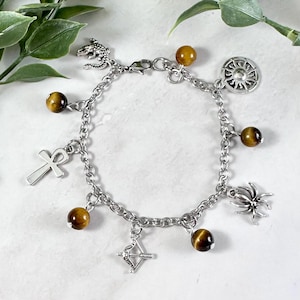 May include: A silver charm bracelet with tiger's eye beads and various charms, including a sun, an ankh, a spider, a lizard, and an arrow.