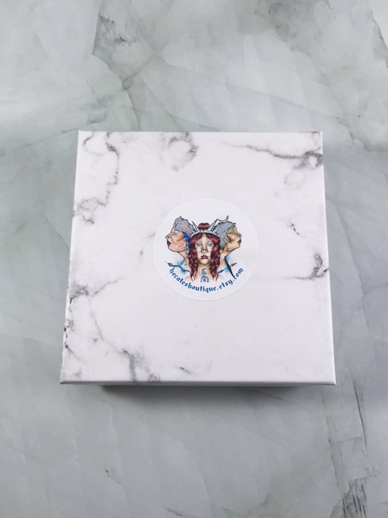 May include: A white marble box with a round sticker featuring a colorful illustration of three women with blue and white clothing. The sticker also includes the text "becatesboutique.etsy.com".