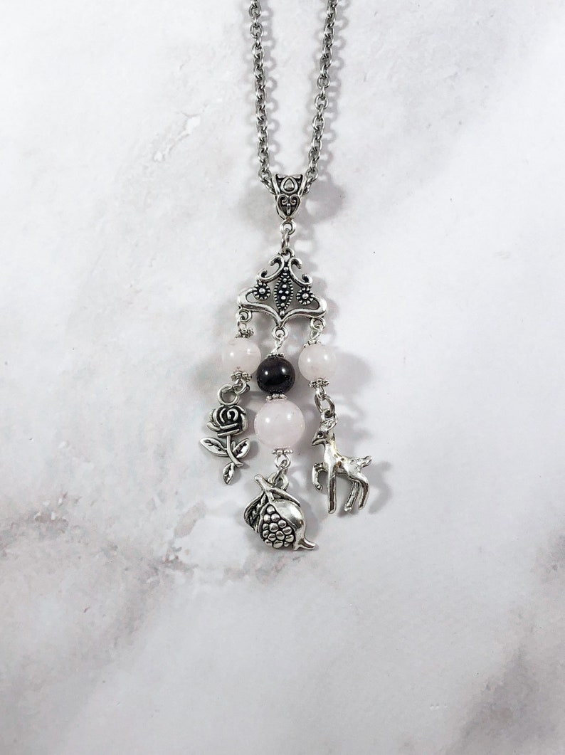 Puede incluir: Un collar de cadena de plata con un colgante de plata que presenta una rosa, un ciervo y una granada. El colgante est&aacute; adornado con cuentas rosas y negras.