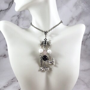 May include: A silver chain necklace with a pendant featuring a crown, three beads, and two silver charms. The beads are white, pink, and black. The charms are a feather and a small animal.