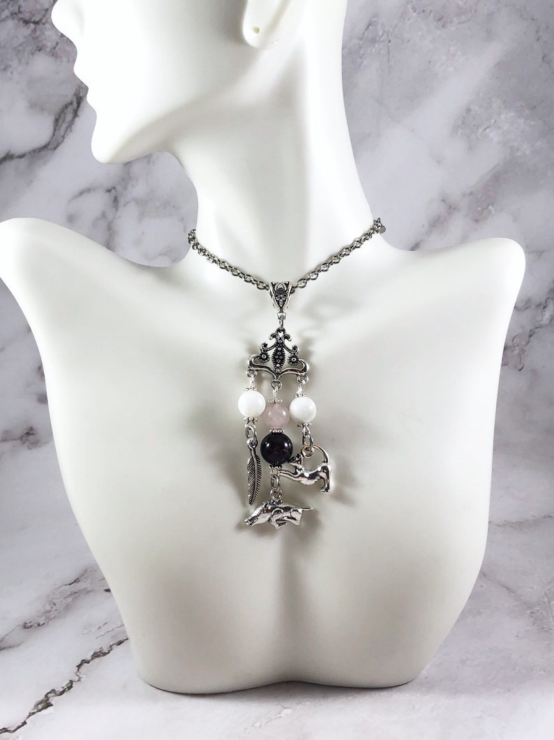 May include: A silver chain necklace with a pendant featuring a crown, three beads, and two silver charms. The beads are white, pink, and black. The charms are a feather and a small animal.