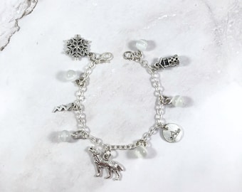 May include: Silver charm bracelet featuring a snowflake, a howling wolf, and other charms. The bracelet includes small, round, white beads and is displayed on a white surface.