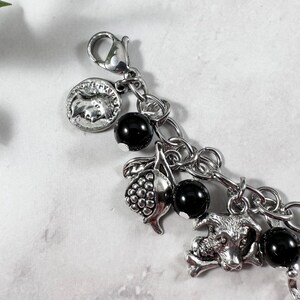 May include: Silver-toned charm bracelet featuring a variety of charms. Charms include a coin, a bunch of grapes, a dog with a bone, and black beads. The bracelet is attached with a lobster clasp.