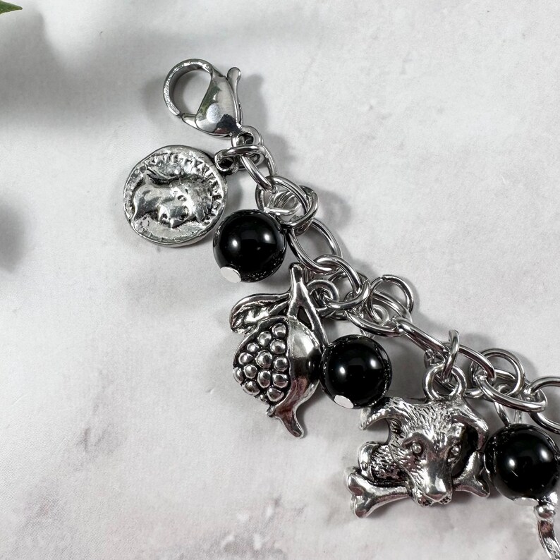 May include: Silver-toned charm bracelet featuring a variety of charms. Charms include a coin, a bunch of grapes, a dog with a bone, and black beads. The bracelet is attached with a lobster clasp.