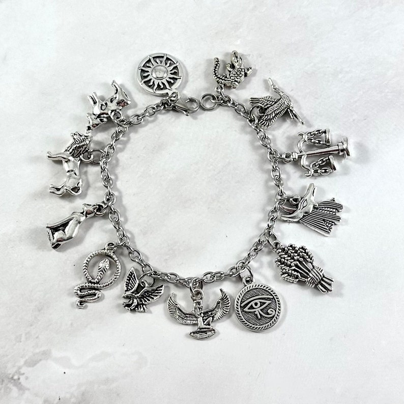 May include: Silver charm bracelet with various Egyptian-themed charms, including a sun, an ankh, a scarab beetle, a cobra, a cat, a lion, a falcon, a scale, a wheat sheaf, and an eye of Horus.