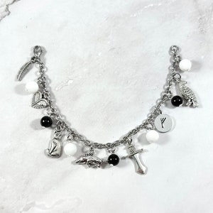 May include: A silver charm bracelet with white and black beads and charms, including a feather, heart, cat, dolphin, sword, raven, and a rune symbol.