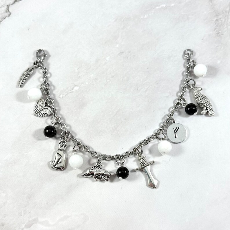 May include: A silver charm bracelet with white and black beads and charms, including a feather, heart, cat, dolphin, sword, raven, and a rune symbol.