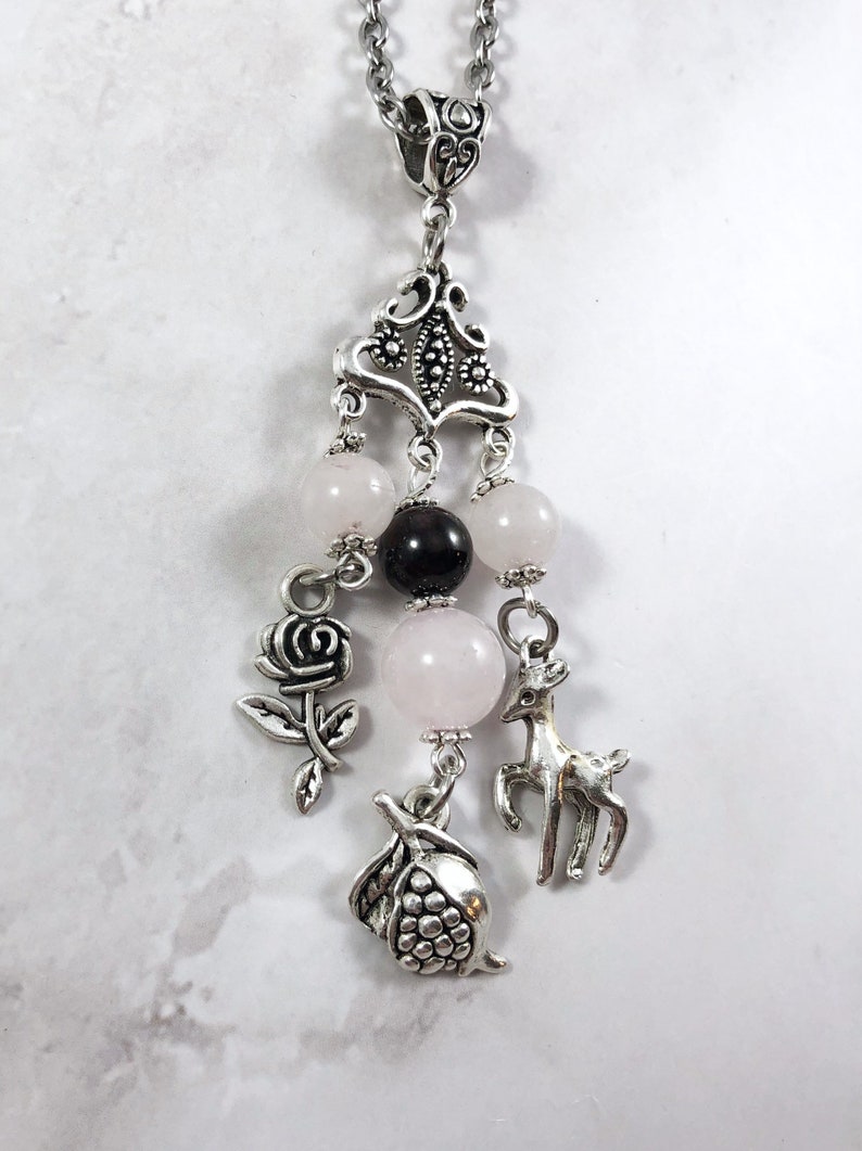 Puede incluir: Un collar con dije de plata con dijes de rosa, ciervo y granada. El collar tambi&eacute;n tiene cuentas rosadas y negras.
