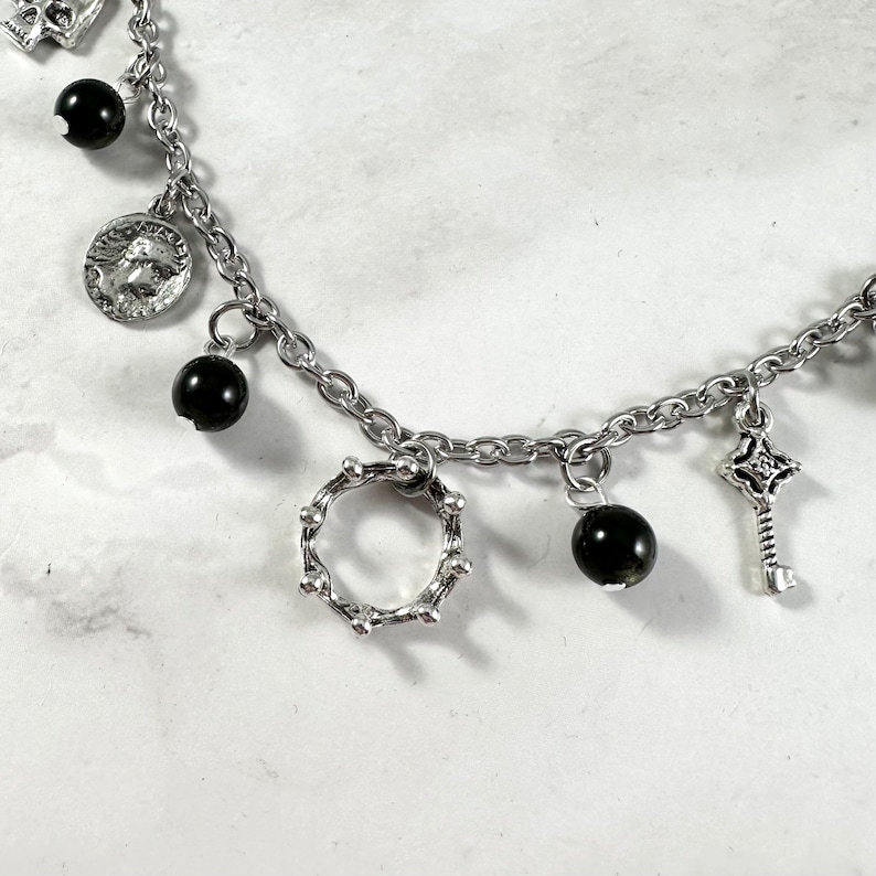 May include: A silver chain bracelet with multiple charms, including a small crown, a key, and black beads.