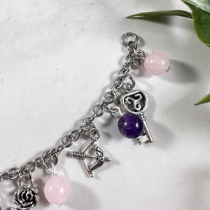 May include: A silver charm bracelet with various charms, including a key, a bow and arrow, and a rose. The bracelet also features pink and purple gemstone beads.