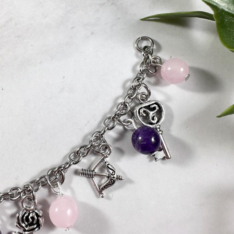 May include: A silver charm bracelet with various charms, including a key, a bow and arrow, and a rose. The bracelet also features pink and purple gemstone beads.