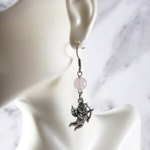 May include: Silver dangle earrings with a pink quartz bead and a silver cherub charm holding a bow and arrow.