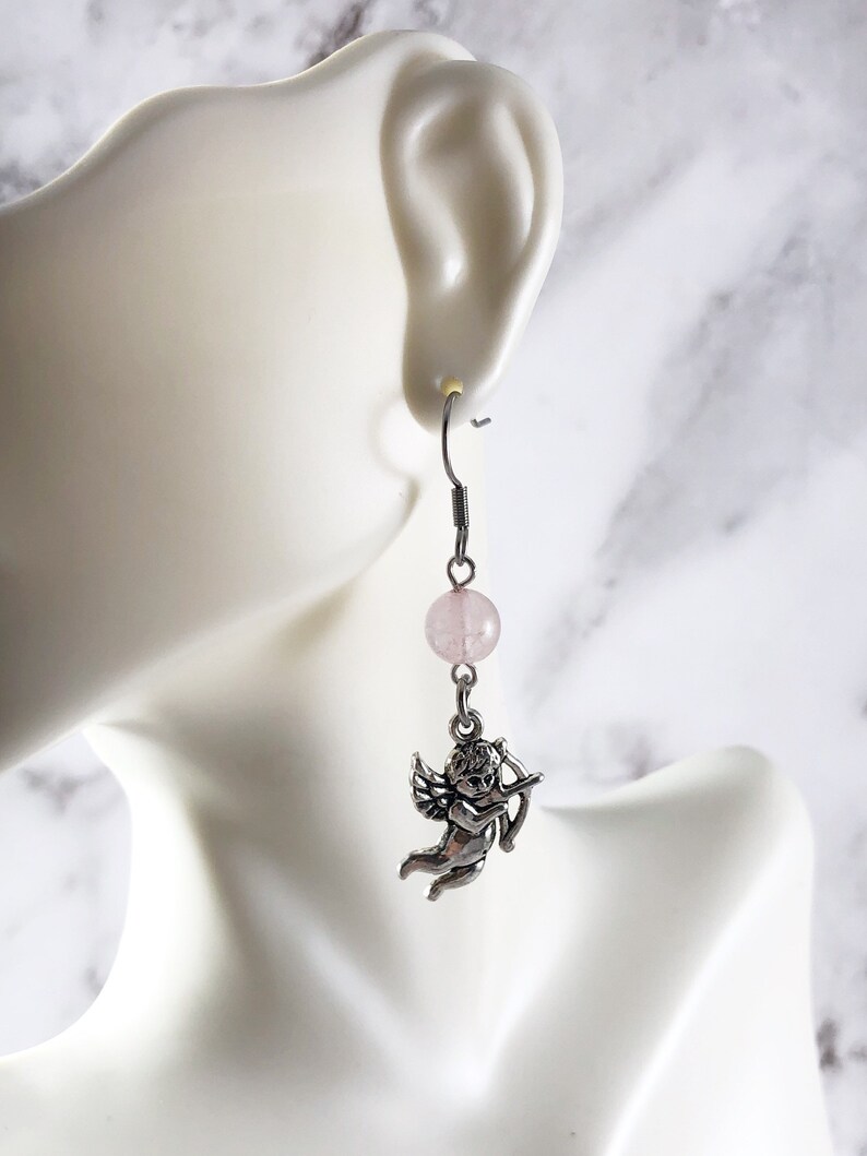 May include: Silver dangle earrings with a pink quartz bead and a silver cherub charm holding a bow and arrow.