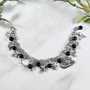 May include: Silver-toned charm bracelet featuring various charms, including skulls, a dragon, and an angel. The bracelet also has black beads interspersed throughout. The bracelet is displayed on a white surface with greenery in the background.