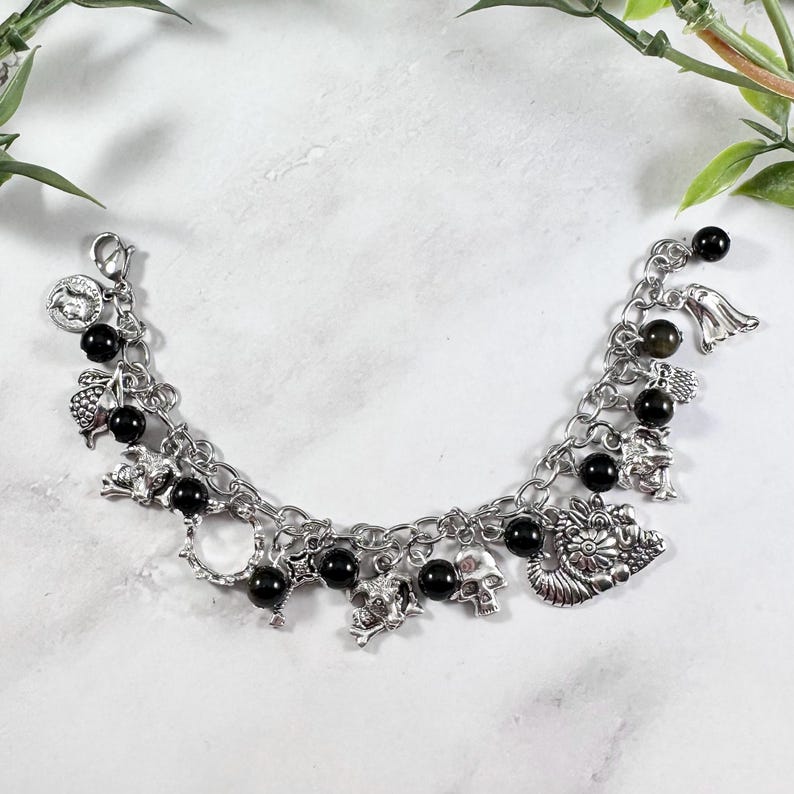 May include: Silver-toned charm bracelet featuring various charms, including skulls, a dragon, and an angel. The bracelet also has black beads interspersed throughout. The bracelet is displayed on a white surface with greenery in the background.