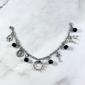 May include: A silver chain bracelet with multiple charms, including a skull, a coin, a crown, a key, and a deer. The charms are interspersed with black beads.