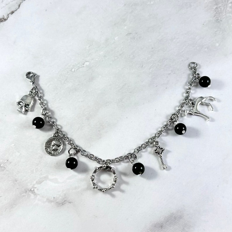 May include: A silver chain bracelet with multiple charms, including a skull, a coin, a crown, a key, and a deer. The charms are interspersed with black beads.