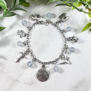 May include: A silver charm bracelet with blue beads and charms, including a labyrinth, a sword, a bull's head, a spool of thread, a trident, a crab, and a Minotaur's head.