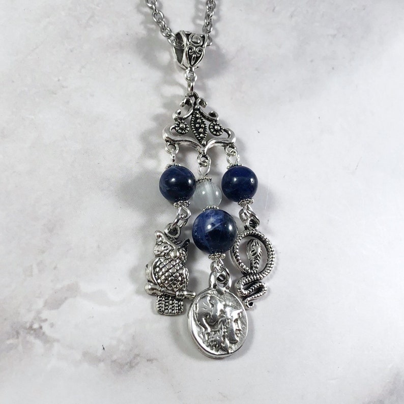 May include: Silver necklace with a blue and white stone pendant. The pendant features an owl, a snake, and a coin charm.