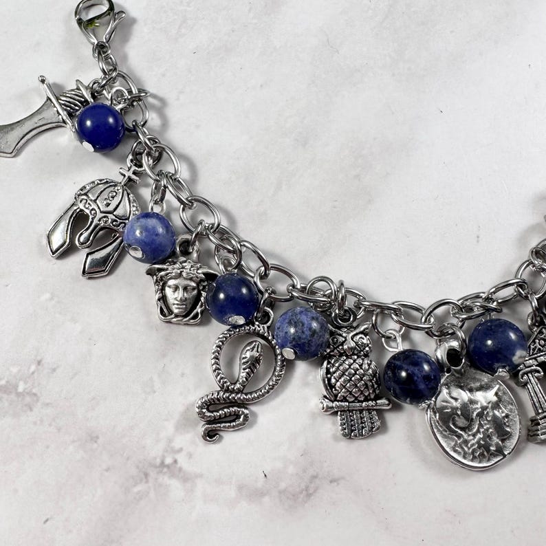 May include: A silver charm bracelet featuring blue beads and a variety of charms, such as a snake, an owl, a Medusa head, and a helmet.