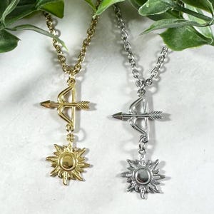 Puede incluir: Dos collares con un colgante de sol y arco y flecha. Un collar tiene un colgante dorado y el otro tiene un colgante plateado.