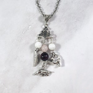 May include: A silver necklace with a charm featuring a pink and white stone, a silver feather, and two silver animal charms. The necklace is on a white background.