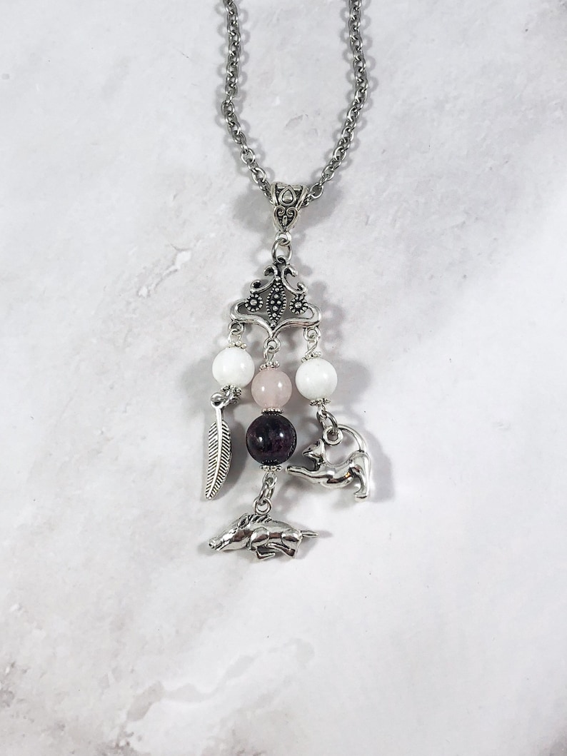 May include: A silver necklace with a charm featuring a pink and white stone, a silver feather, and two silver animal charms. The necklace is on a white background.