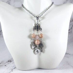 May include: A silver chain necklace with a pendant featuring a silver crown, pink and white beads, a silver swan charm, a silver wheat charm, and a silver charm with the rune "Algiz" engraved on it.
