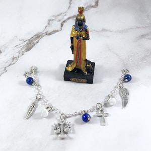 May include: A silver charm bracelet with blue beads, white beads, and silver charms including a feather, scales of justice, an ankh, and a wing. The bracelet is on a white marble surface with a small statue of Maat, the Egyptian goddess of truth and justice.