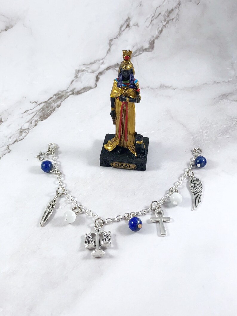 May include: A silver charm bracelet with blue beads, white beads, and silver charms including a feather, scales of justice, an ankh, and a wing. The bracelet is on a white marble surface with a small statue of Maat, the Egyptian goddess of truth and justice.