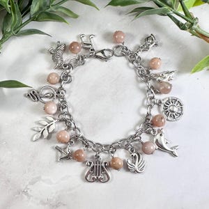 Op de afbeelding: Een zilverkleurige bedelarmband met een reeks ronde, licht perzikkleurige kralen afgewisseld met diverse metalen bedels, waaronder een zon, een dolfijn, een zwaan, een lier, een pijl en boog, een bladerige tak, een slang en een muzieknoot.