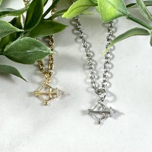 May include: Two silver necklaces with a bow and arrow pendant. One necklace has a gold chain and the other has a silver chain.
