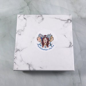 May include: White marble gift box with a round sticker featuring a colorful illustration of three faces and the text "hecatesboutique.etsy.com".