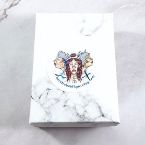 May include: White box with a round sticker featuring a colorful illustration of three faces with a crown and the text "becatesboutique.etsy.com".