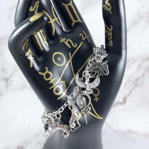 May include: Silver charm bracelet with multiple charms, including an ankh, eye of Horus, snake, lion, and cat. The bracelet is on a black hand with gold writing that says "will logic".