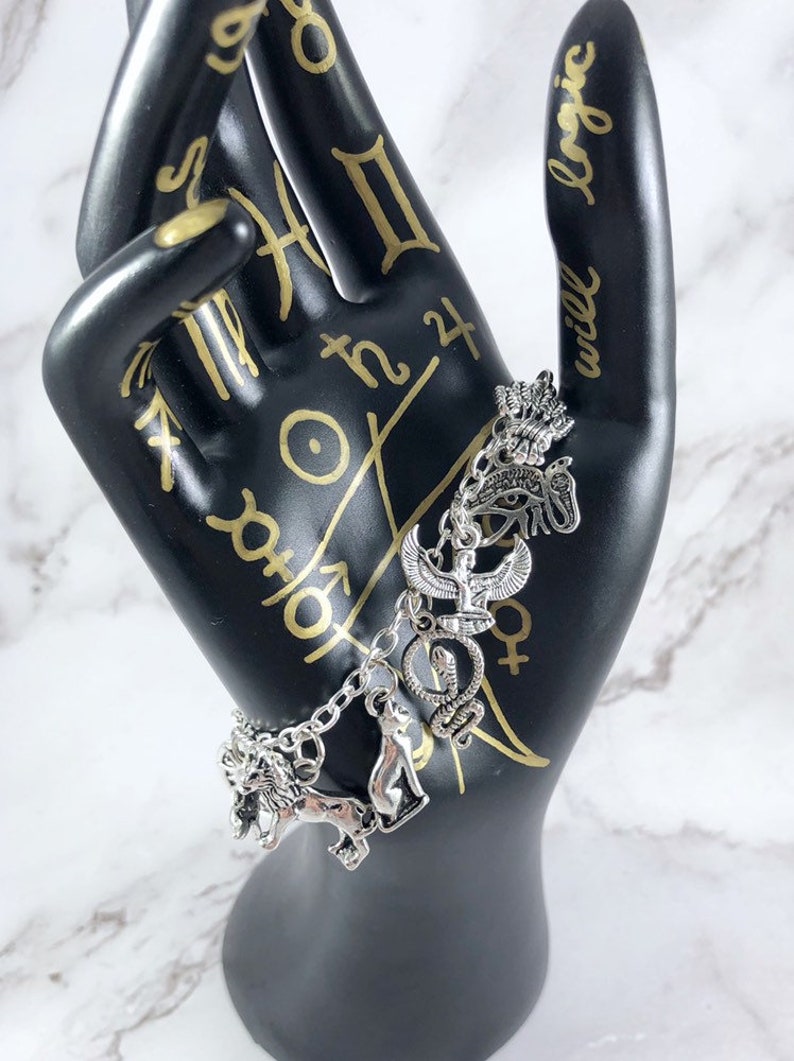 May include: Silver charm bracelet with multiple charms, including an ankh, eye of Horus, snake, lion, and cat. The bracelet is on a black hand with gold writing that says "will logic".
